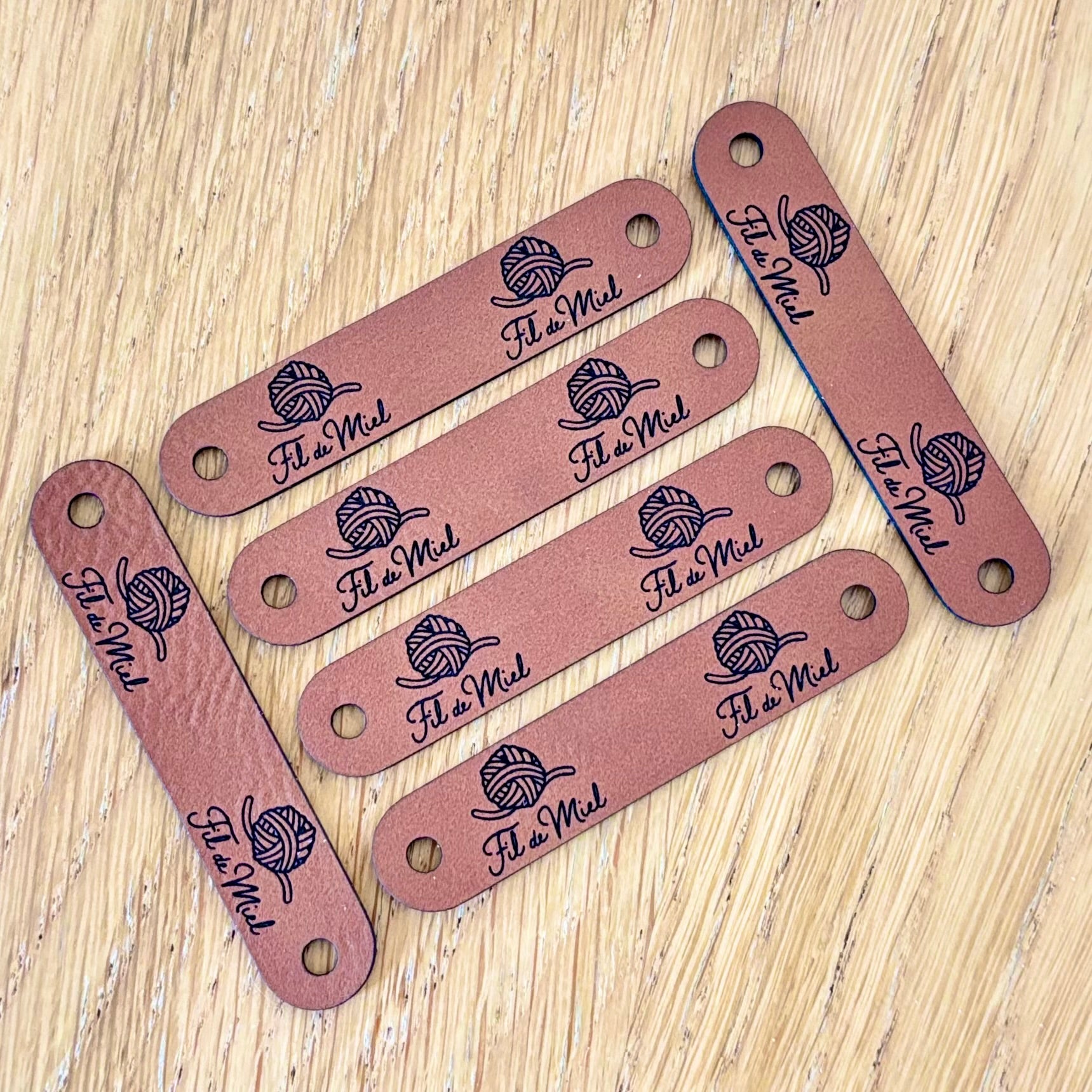 Etiquettes de finition en simili cuir couleur rose, imprimées « Fil de Miel » avec logo boule de fil et petit troue-hole pour languette.