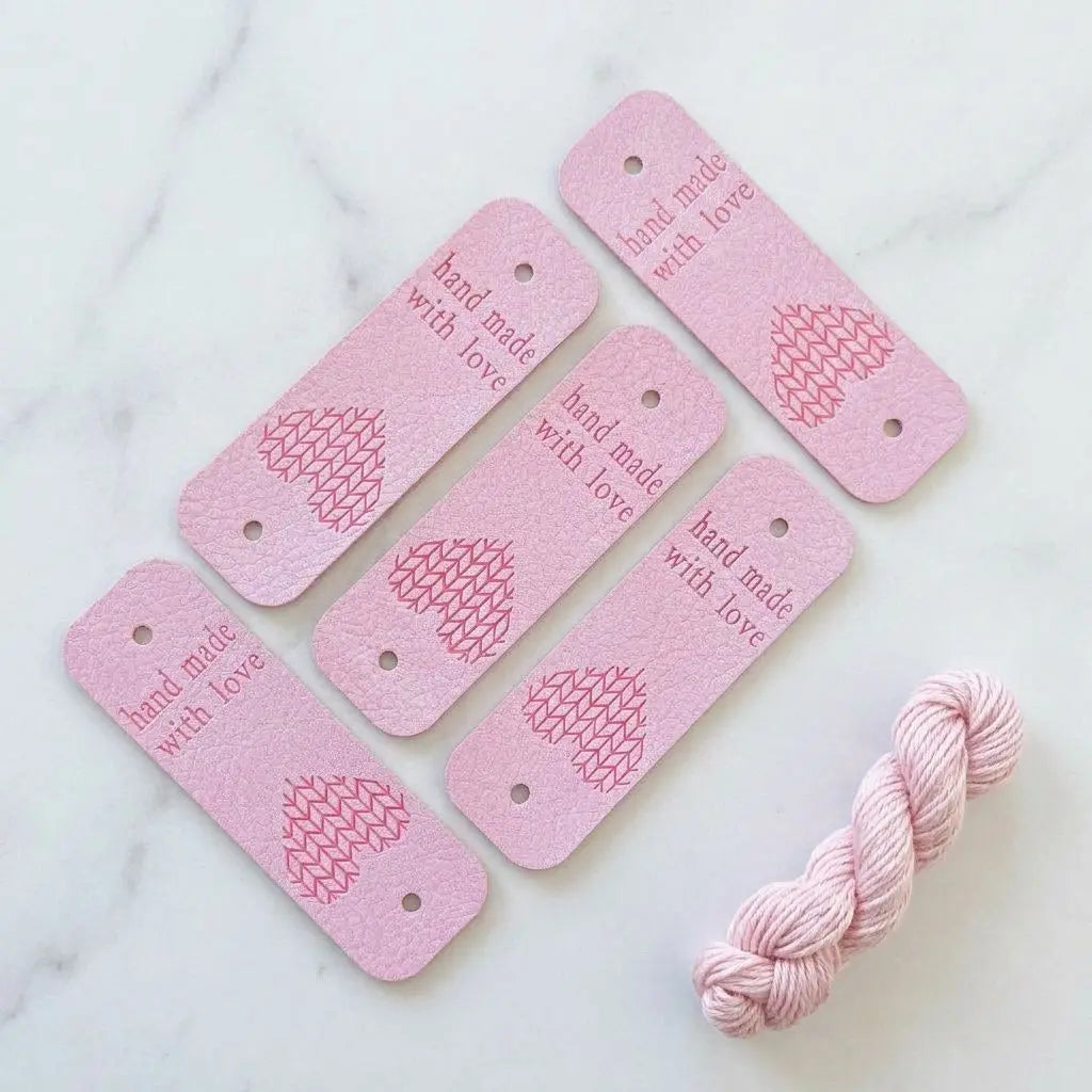 Etiquettes de finition en simili cuir rose, imprimées "hand made with love" avec motif en forme de cœur et petit trou de fixation.