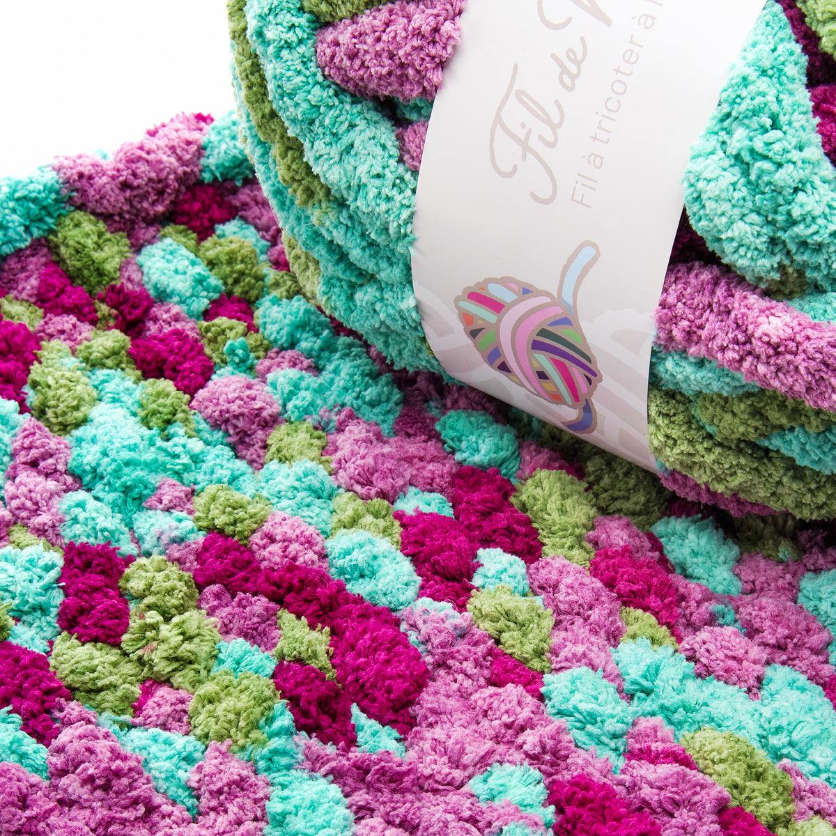 Tapis pelucheux multicolore en forme de Pompon, motifs zébrés vert, turquoise, magenta et violet; étiquette blanche sur le coté indiquant "Forêt Tropicale".