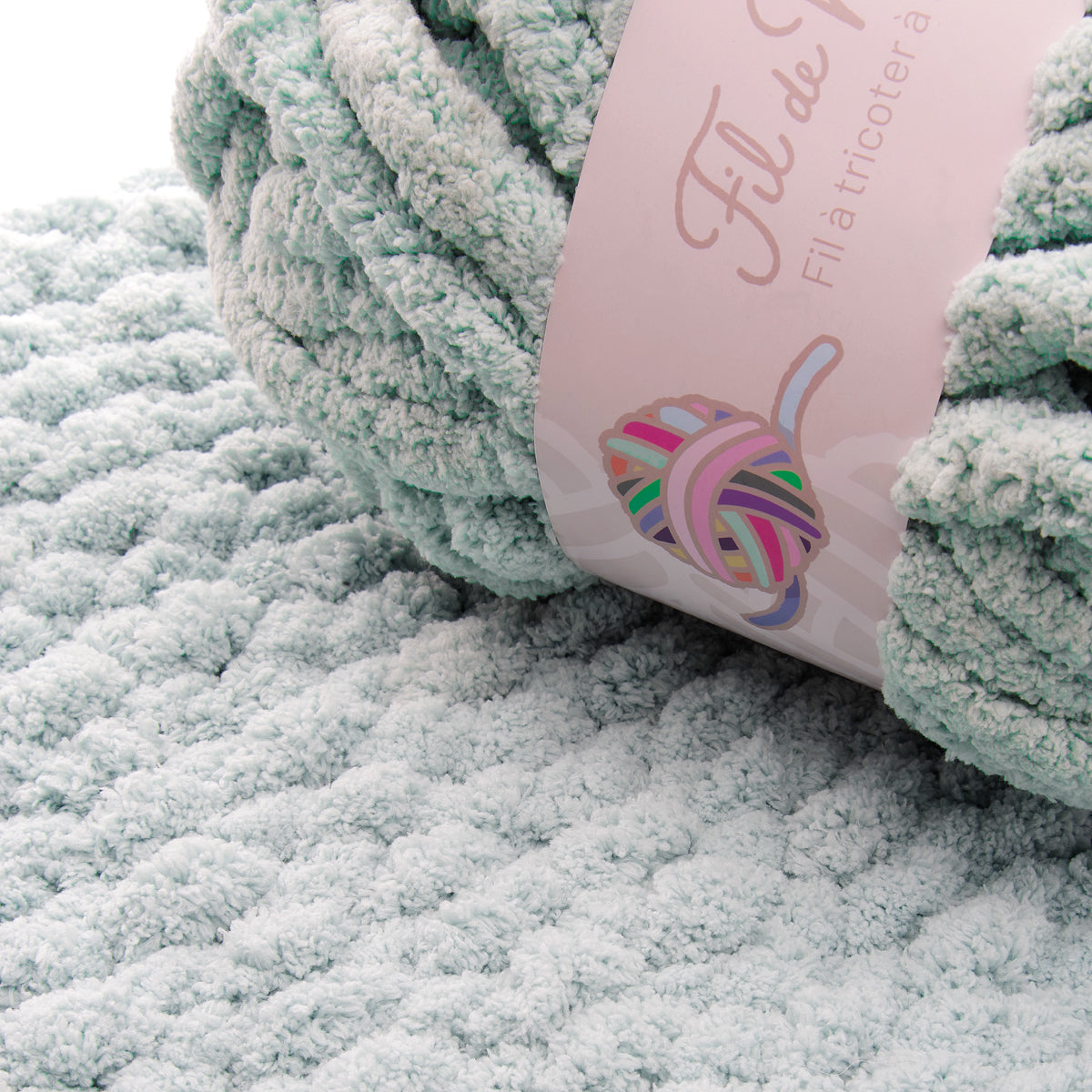 Tricot doux vert d’eau avec étiquette rose affichant un motif de pelote de laine multicolore.