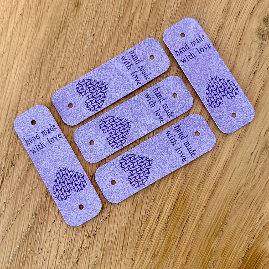 Étiquettes décoratives en simili cuir lilas, rectangulaires avec coin arrondi, motif géométrique en noir et texte « hand made with love », petit trou pour ficelle.