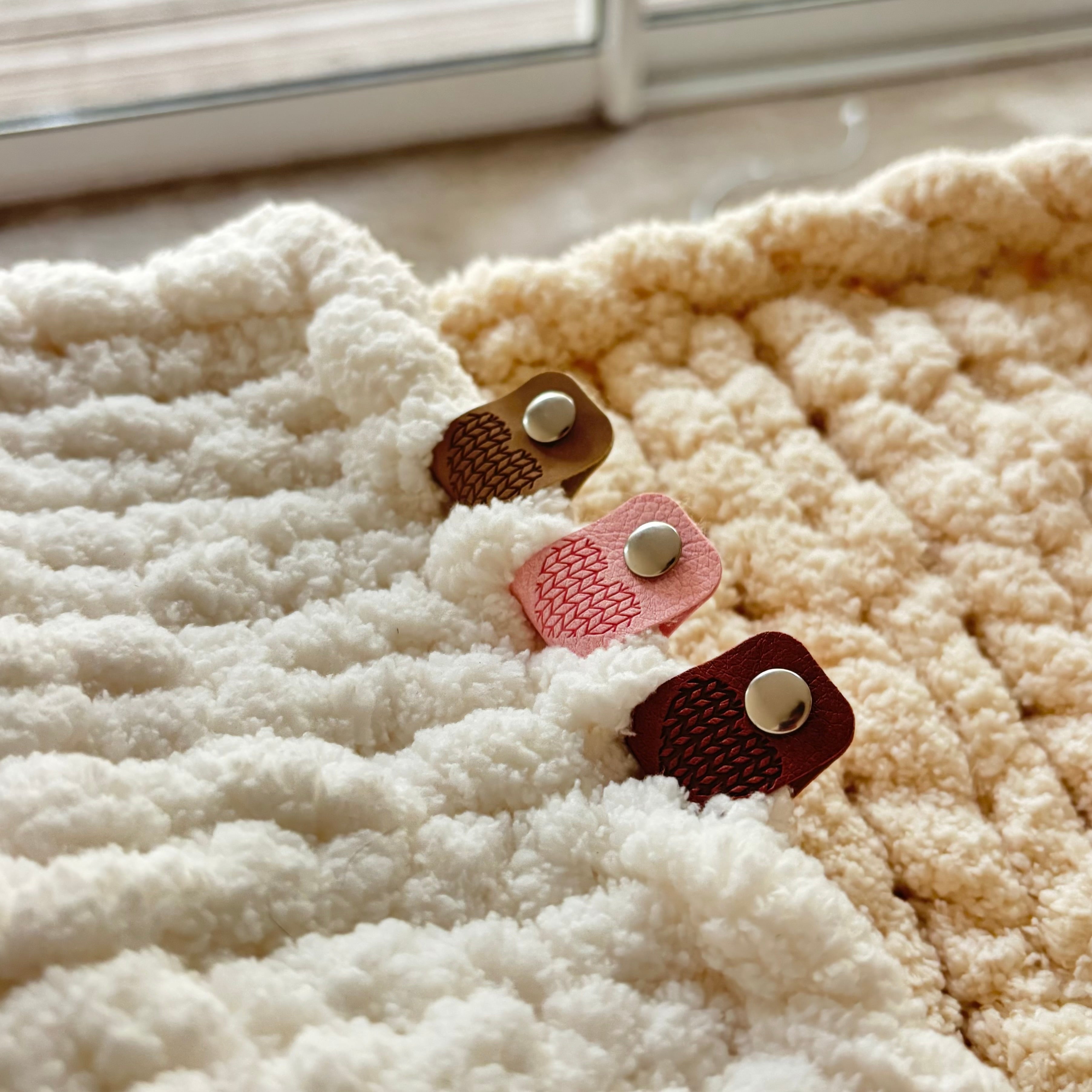 Étiquette de finition en simili cuir avec motif en forme de cœur, rivet métallique et petites anneaux, en brun, rose et rouge sur texture peluche blanche.