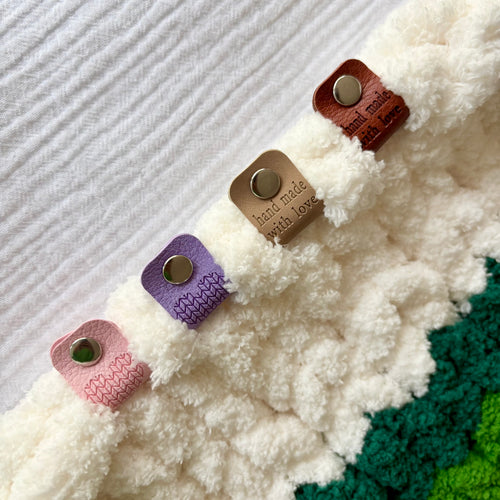 Étiquette de finition en simili cuir de différentes couleurs (rose, beige, violet, brun) fixée sur une peluche blanche avec bouton métallique.