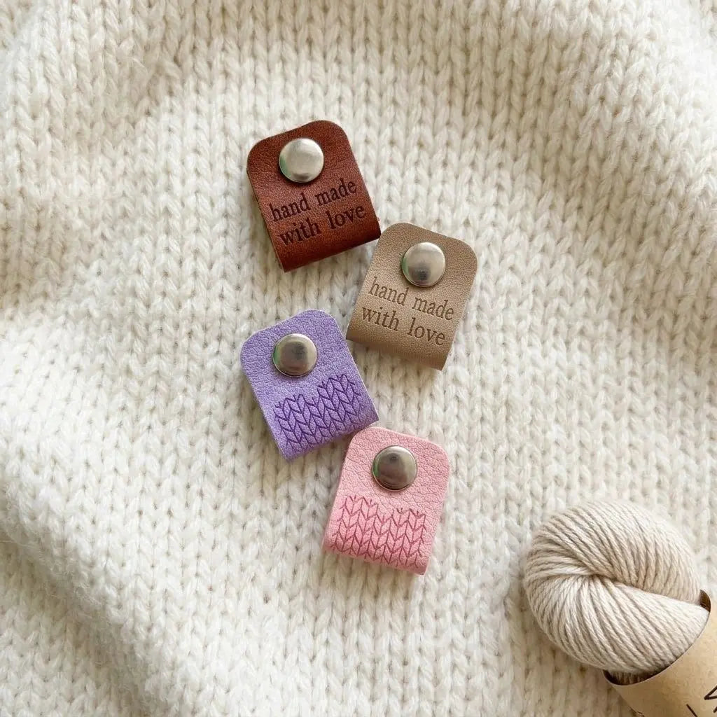Étiquette de finition en simili cuir, multicolore avec bouton métallique sur tricot blanc, texte "hand made with love" sur chaque étiquette.